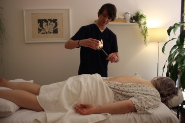 Person lying on a bed with a patterned blanket, another person standing nearby holding a small object, room with artwork and plants