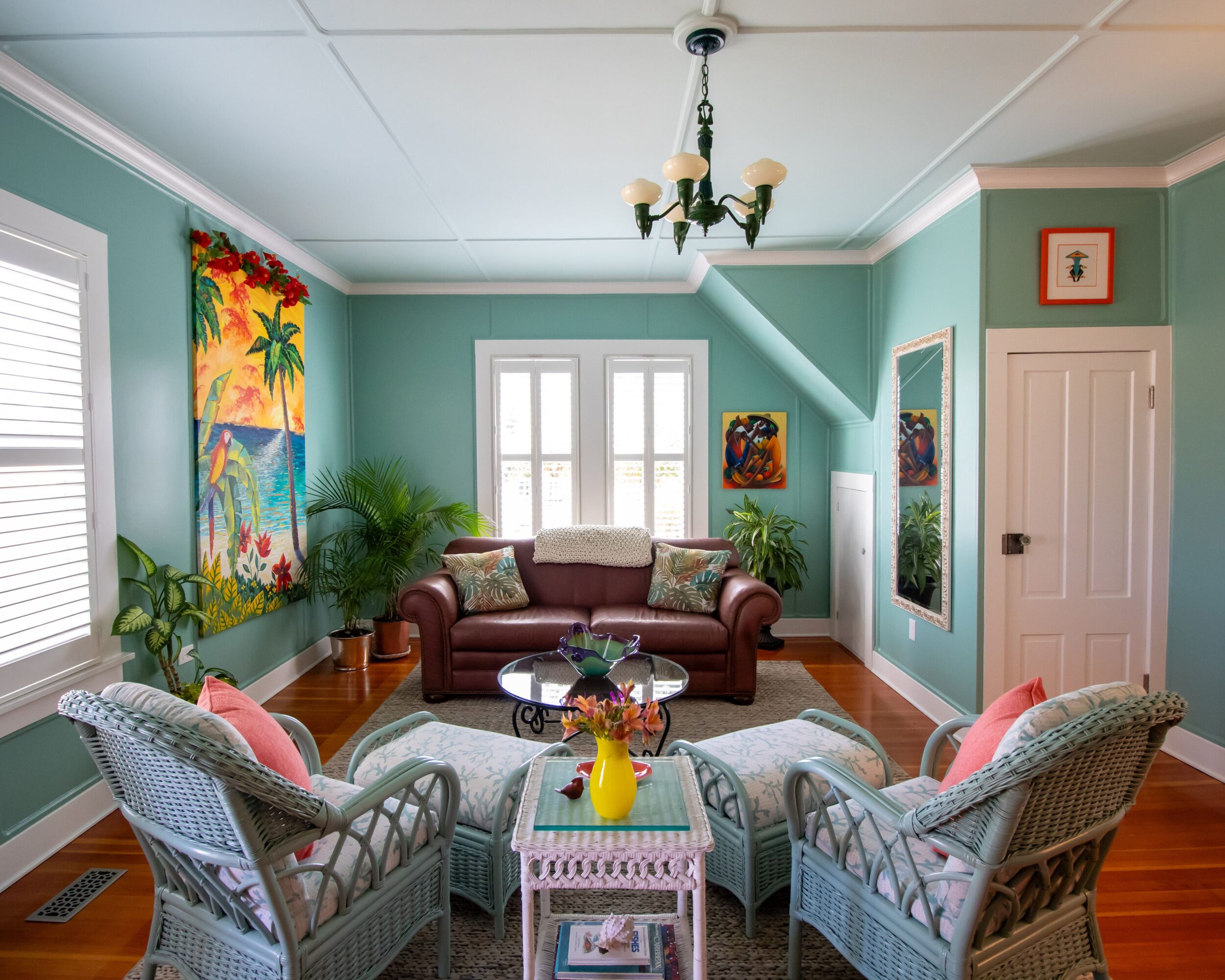 Living room with a brown sofa, two wicker chairs, colorful artwork, and a chandelier, with windows and plants.