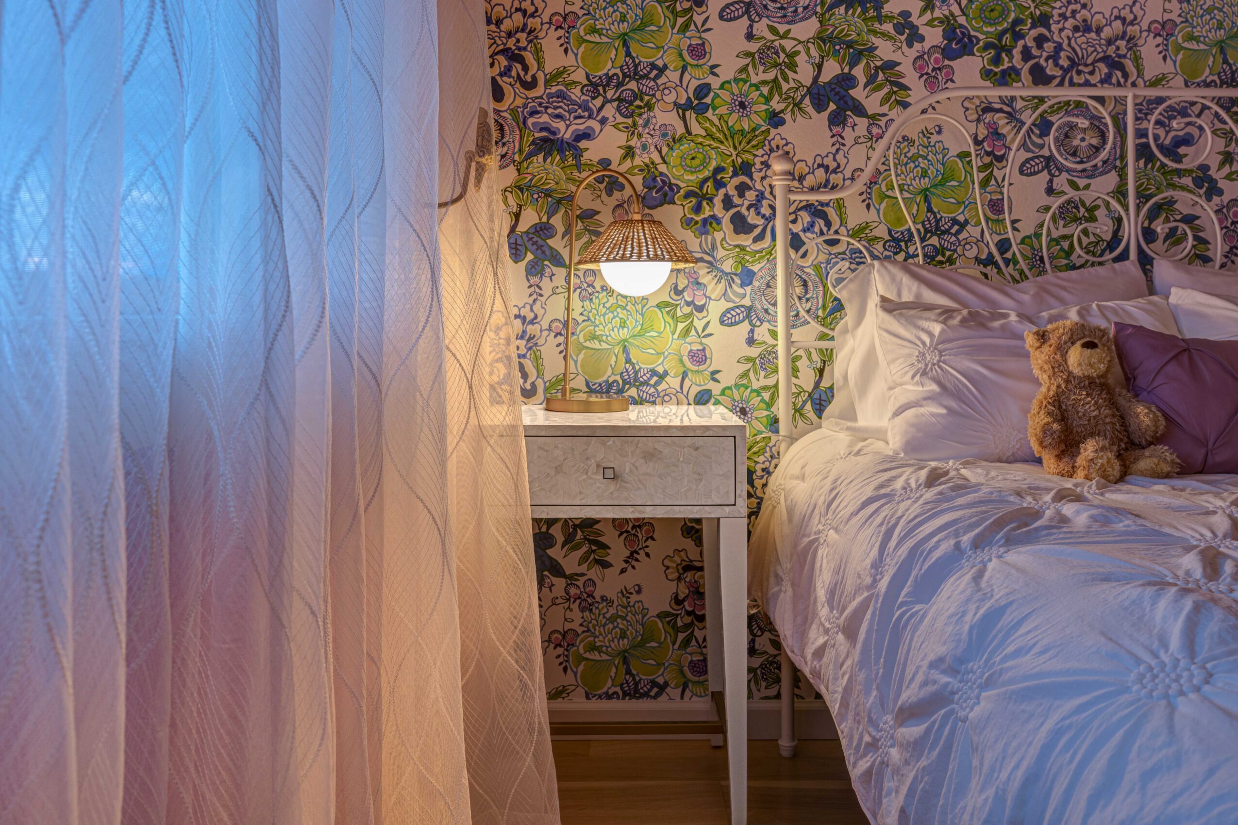 Bedroom with floral wallpaper, bed with white bedding, teddy bear, nightstand, and sheer curtains, illuminated by soft light.