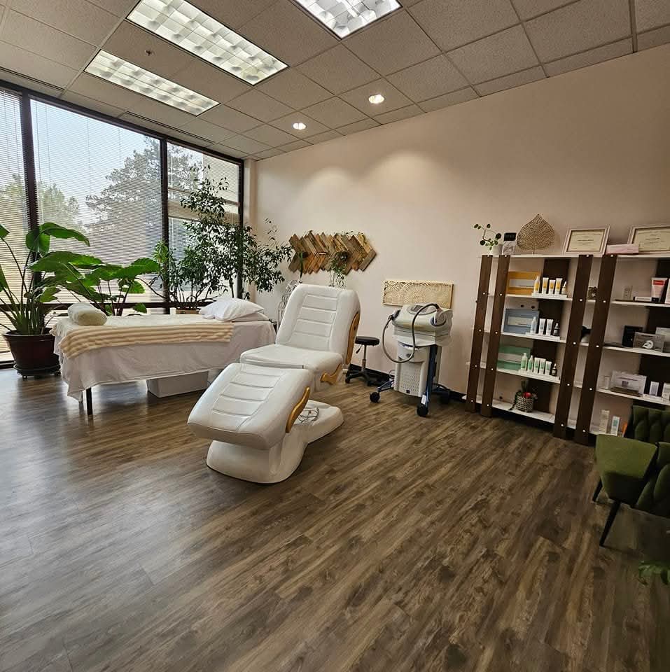 Medical or spa treatment room with a reclining chair, treatment bed, shelves with products, and large window with plants outside.