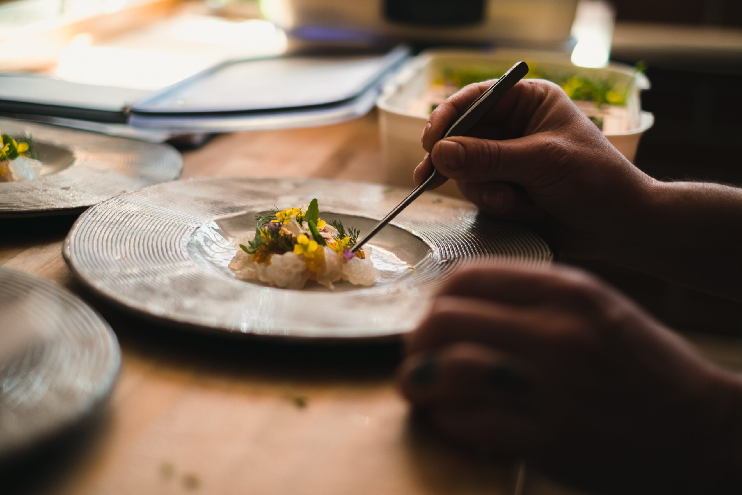 Person using a fork to eat food on a decorative plate, with a blurred background of books and a table.