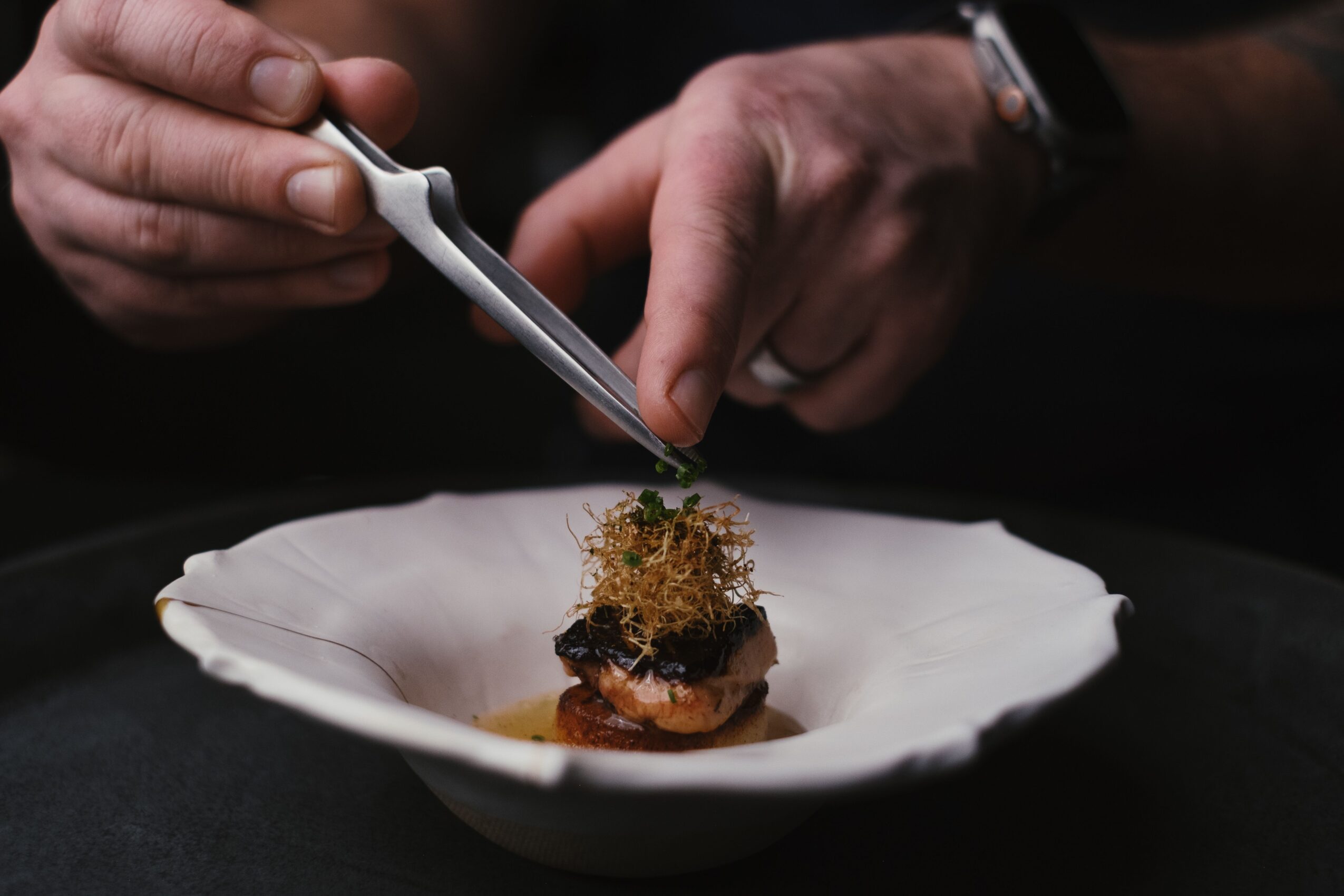 Hands using tweezers to place garnish on a small dish with food, on a dark background.
