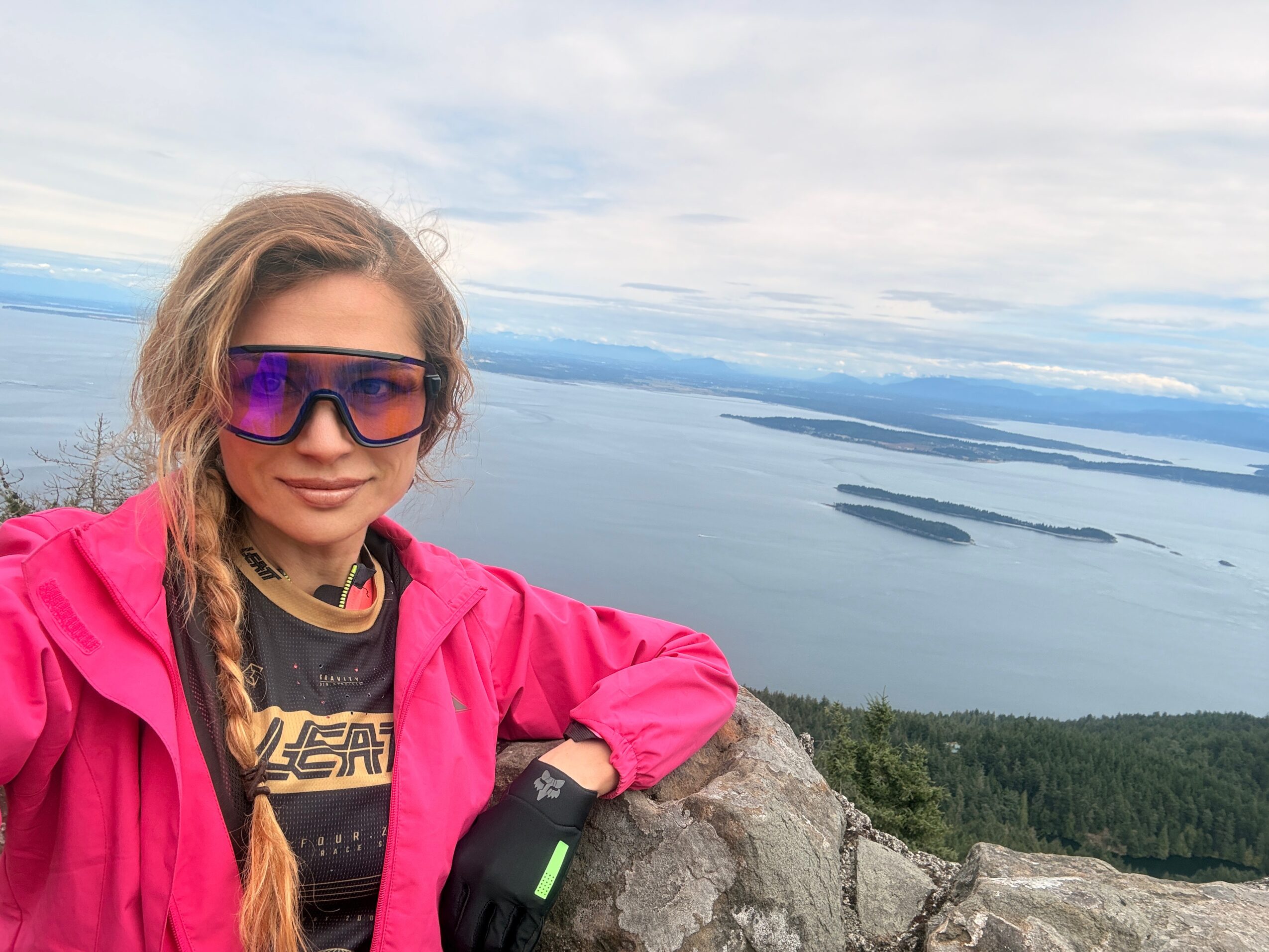 Young woman with long hair in a braid, wearing sunglasses and a pink jacket, standing on a rocky hilltop with a lake view.