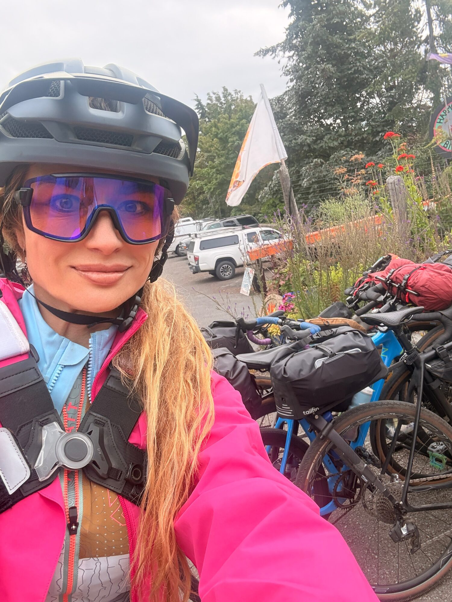 Person wearing a helmet and sunglasses, smiling outdoors with bicycles and a flag in the background.
