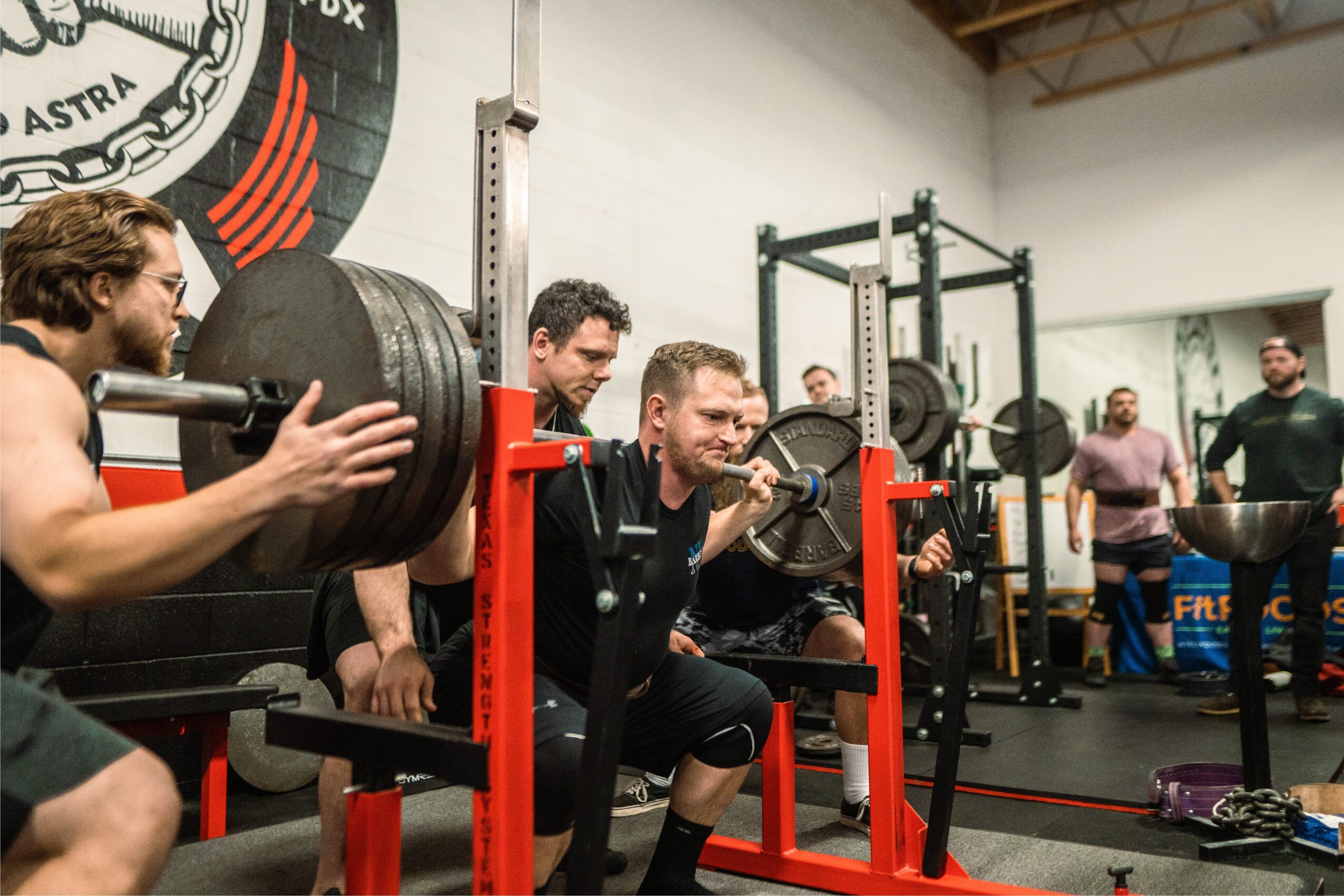 People lifting weights and exercising in a gym with weightlifting equipment and a group of onlookers.