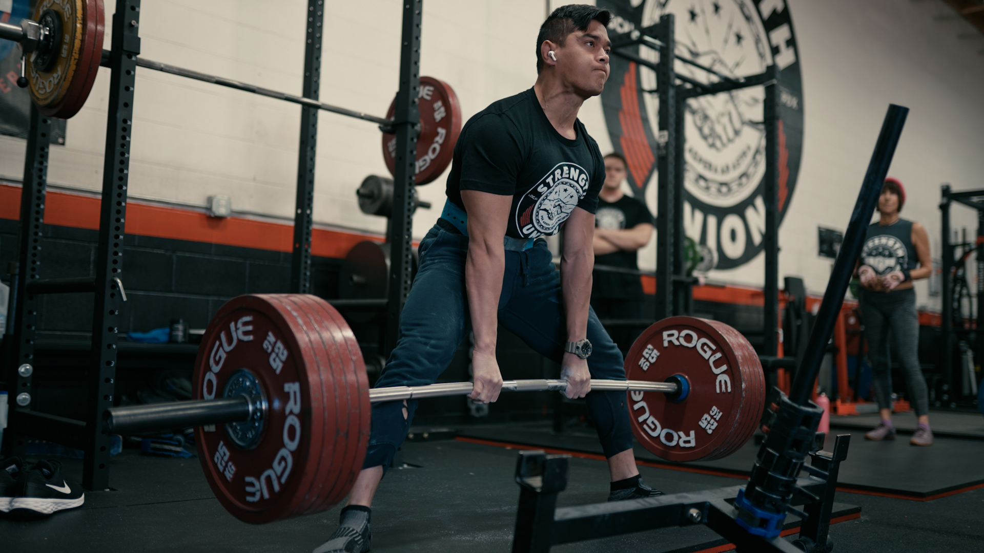 Person lifting a barbell with red weight plates in a gym, others in the background, industrial-style setting.