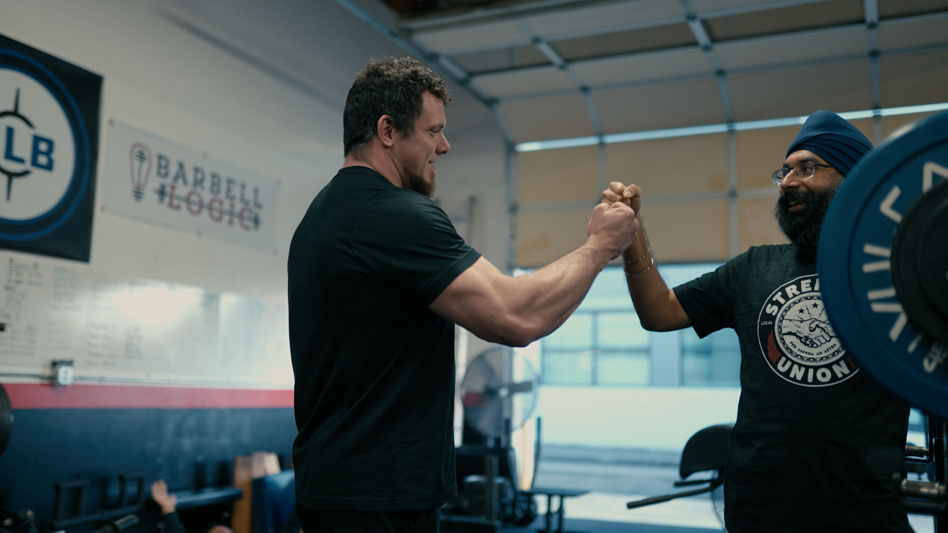 Two men shake hands in a gym, one wearing a black shirt and the other a black T-shirt with a logo.