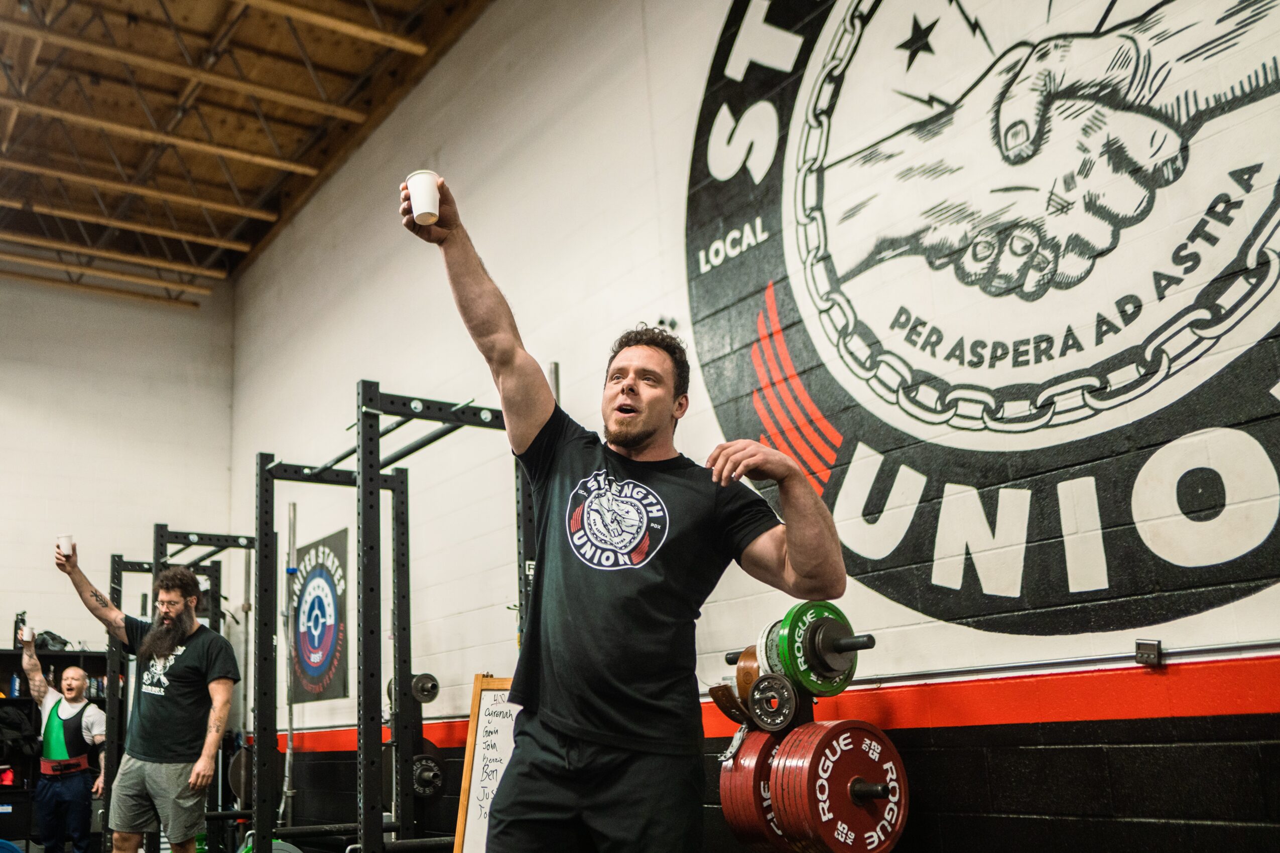 Man lifting a dumbbell in a gym with a large logo on the wall behind him, other people working out in the background.