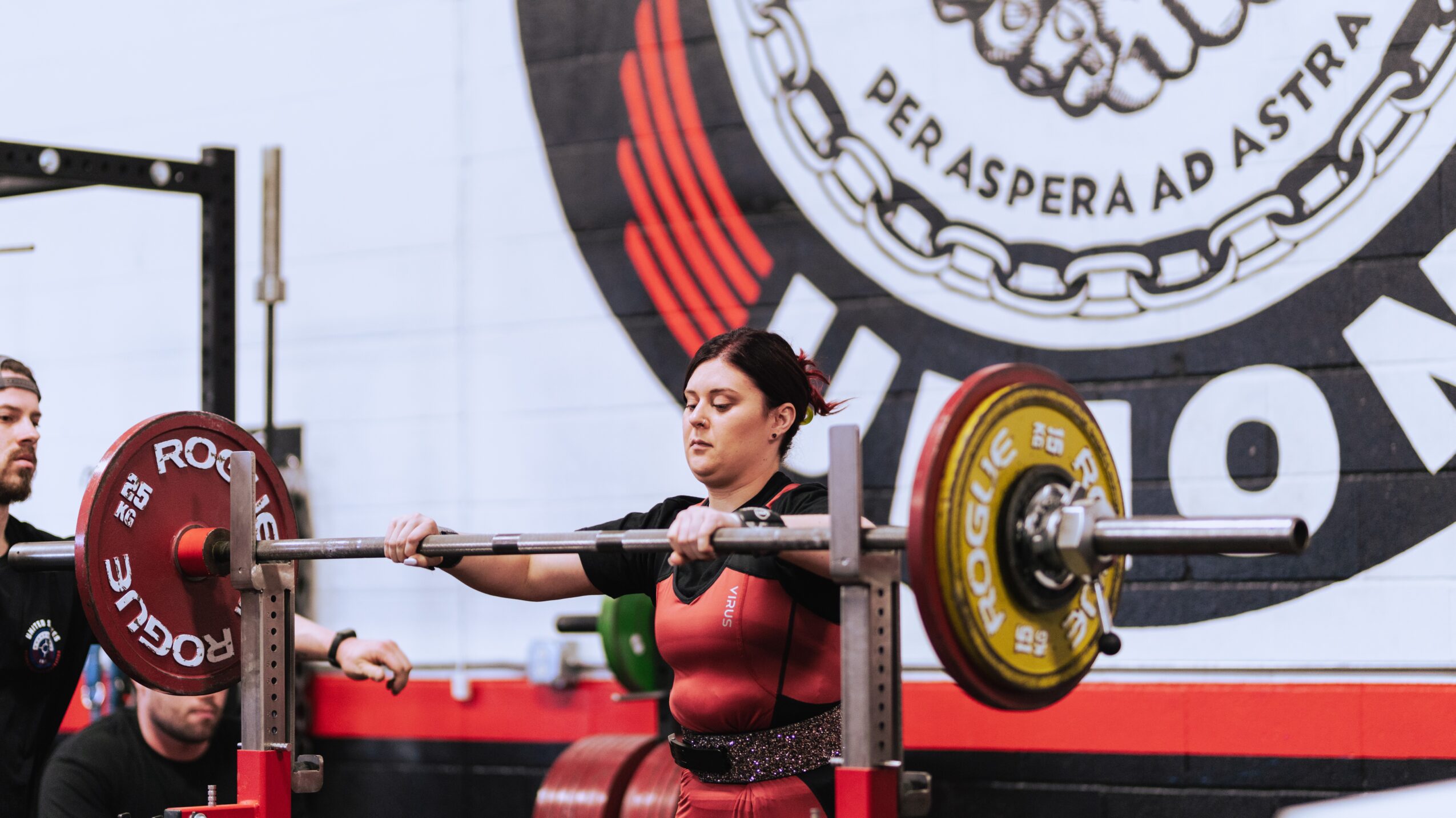 Woman lifting a barbell during a weightlifting competition, with a large circular logo in the background.