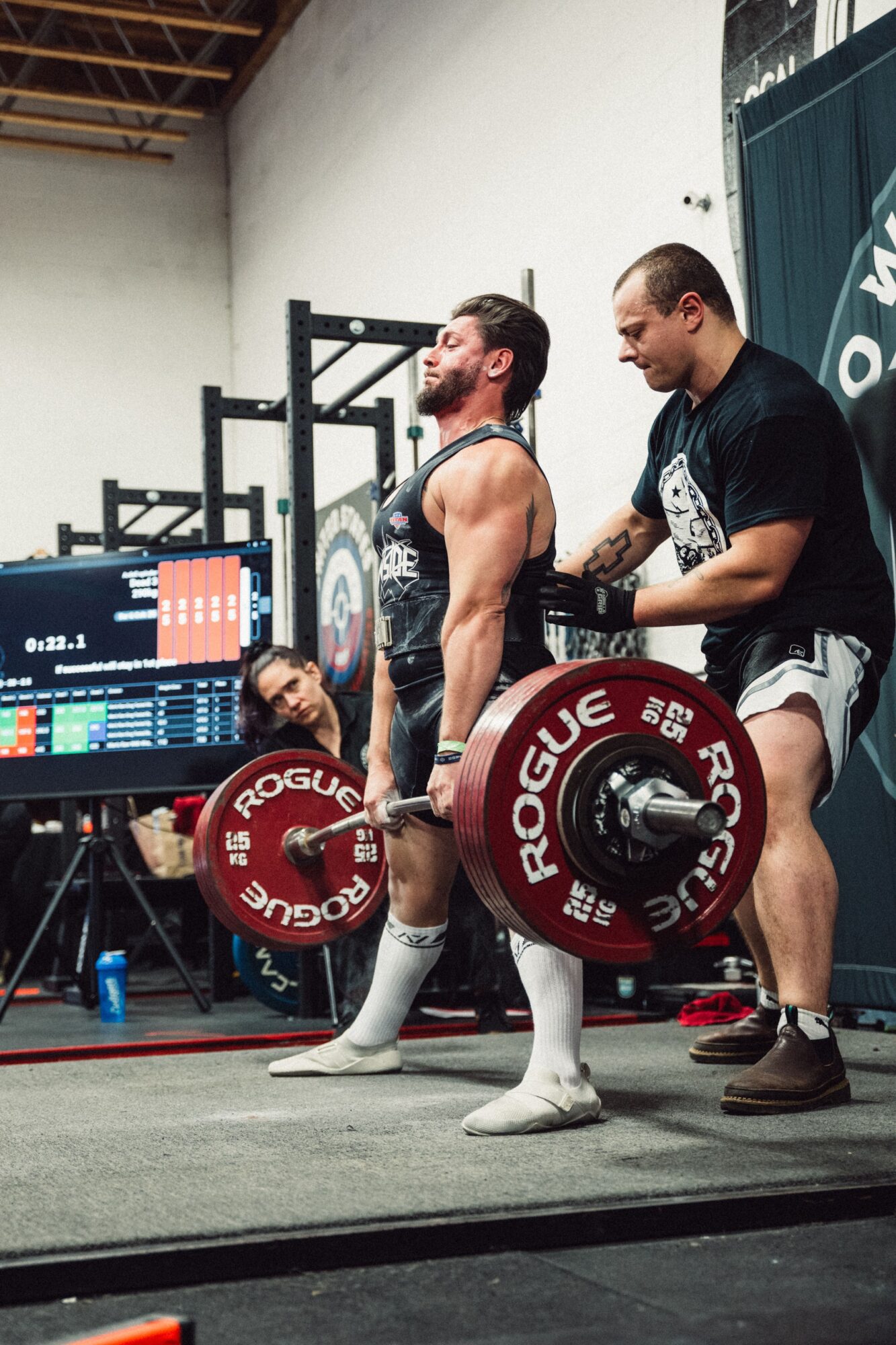 A man lifts a barbell with red Rogue weight plates while a trainer spots him in a gym. A woman watches nearby.