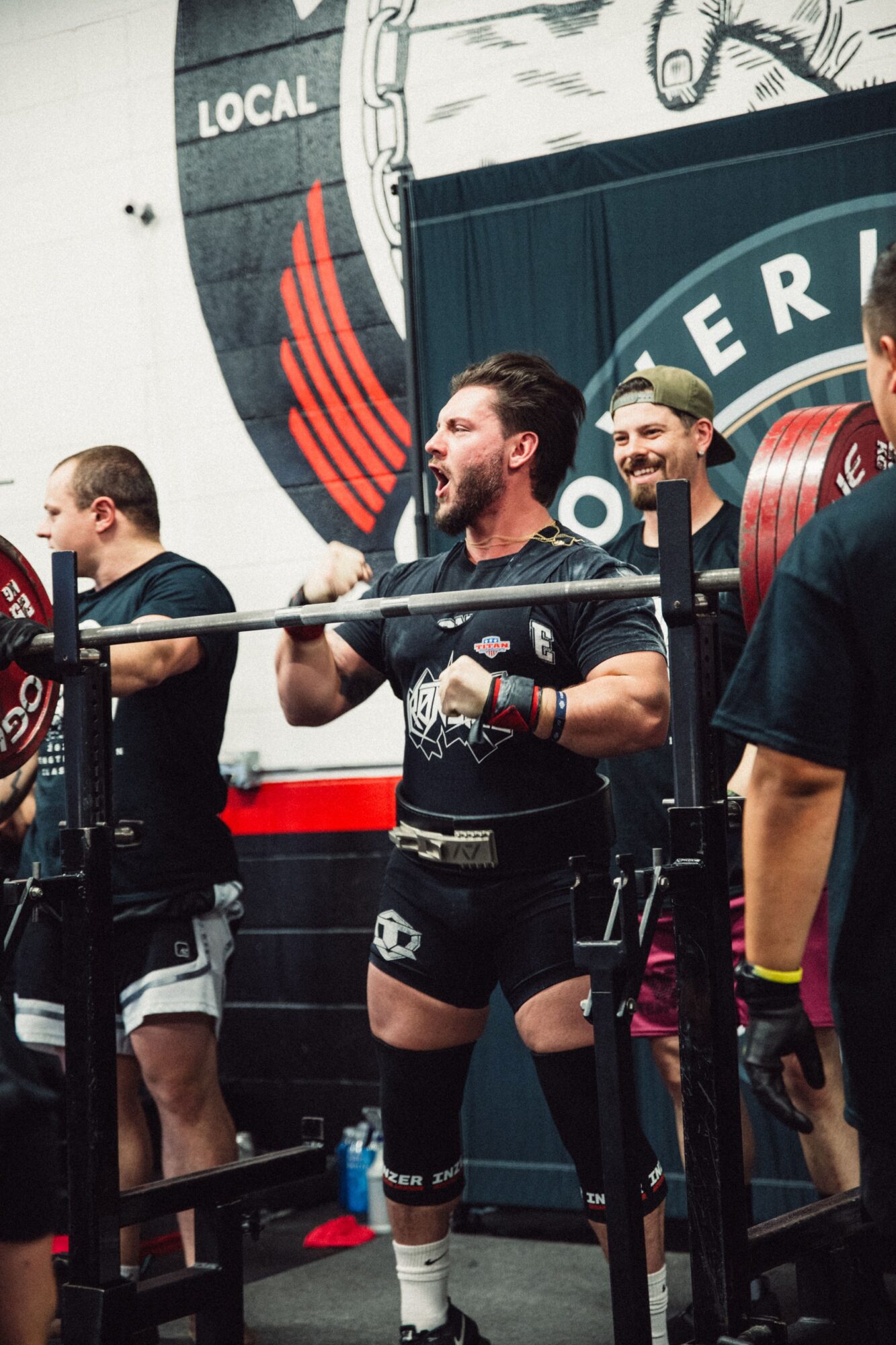 Group of people in a gym, lifting weights and smiling, with a large logo in the background.