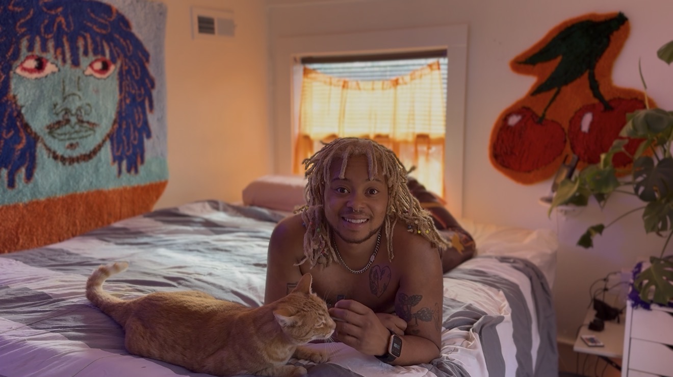 Child with curly hair lying on bed, smiling, with a cat nearby, in a cozy room with colorful wall art and a window.
