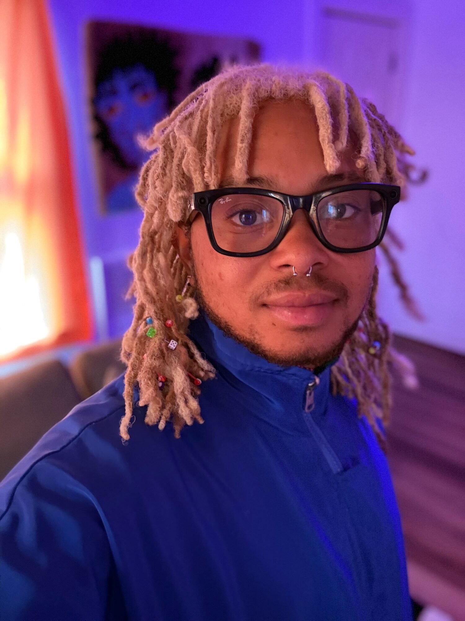 Person with blonde dreadlocks, glasses, and a blue jacket indoors, with a colorful painting in the background.