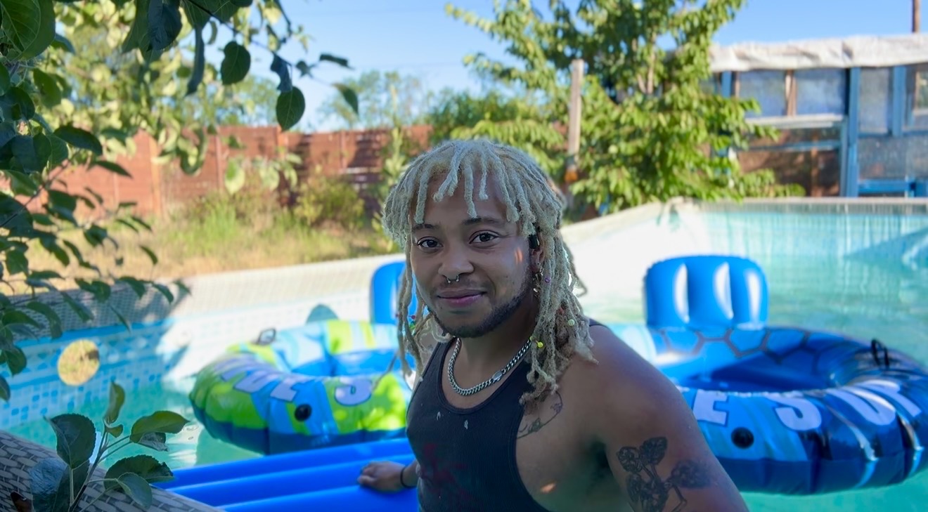 Young man with long, light-colored dreadlocks standing near a pool with inflatable floats, outdoors on a sunny day.