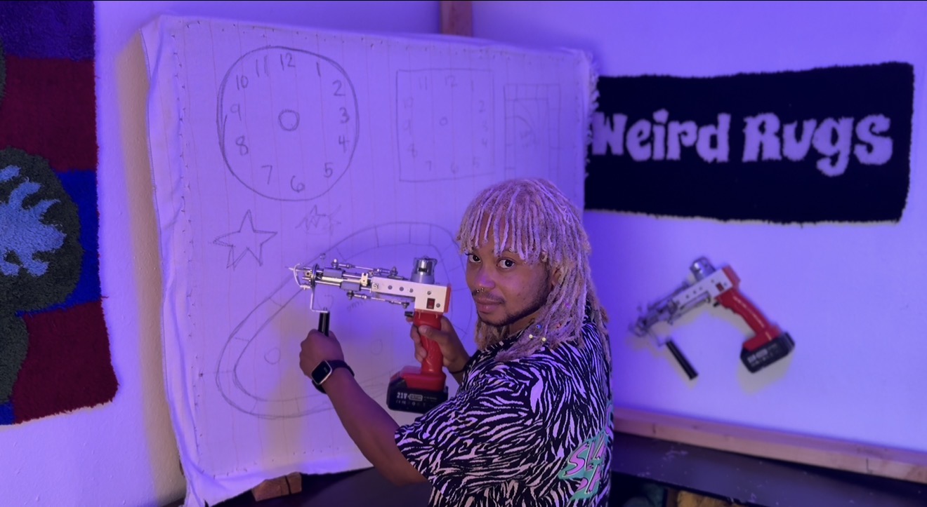 Person with long, light-colored hair holding a toy gun in front of a large notebook with drawings, next to a sign that says 'Weird Rugs'.
