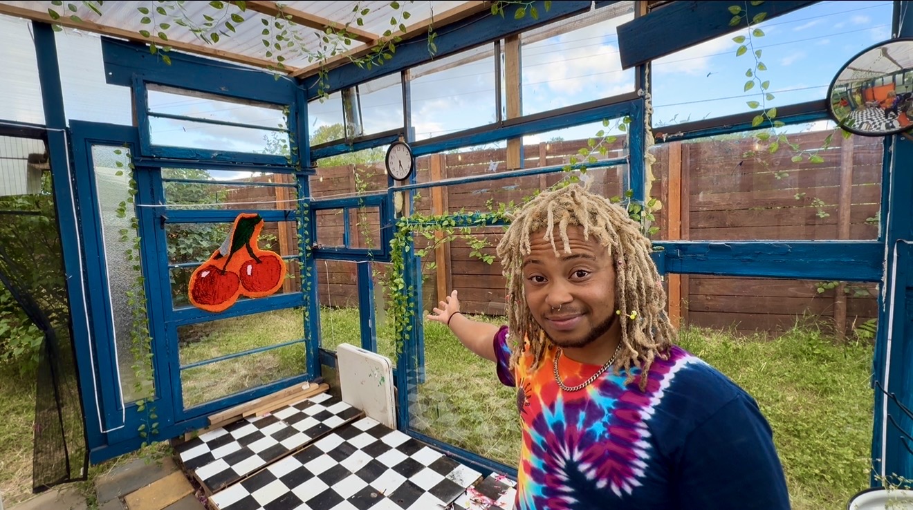 Child with dreadlocks smiling inside a colorful outdoor playhouse with checkered floor, blue walls, and hanging decorations.