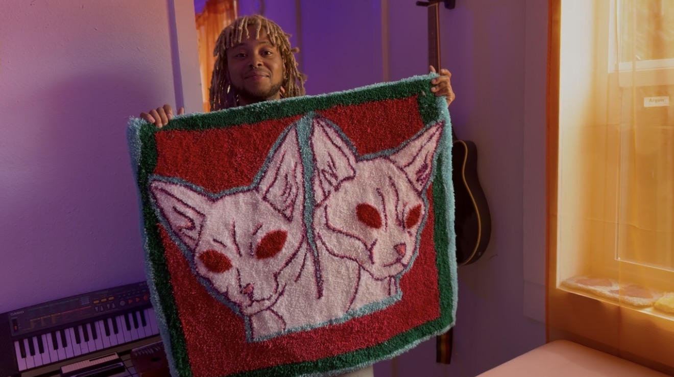 Child holding a colorful blanket with two stylized cat faces, indoors with purple and orange walls.