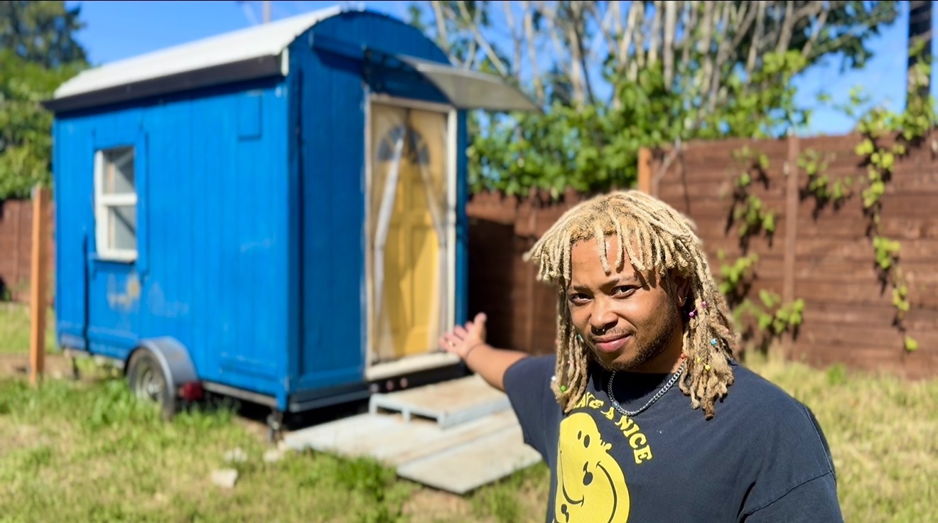 Person with blonde dreadlocks standing outside a small blue shed with a yellow door, in a garden with a wooden fence.