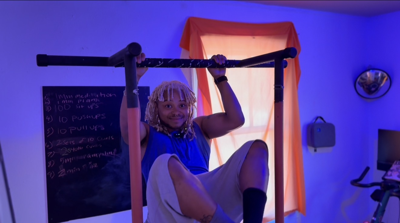 Child hanging from a horizontal bar indoors, with a blackboard and window with orange curtains in the background.