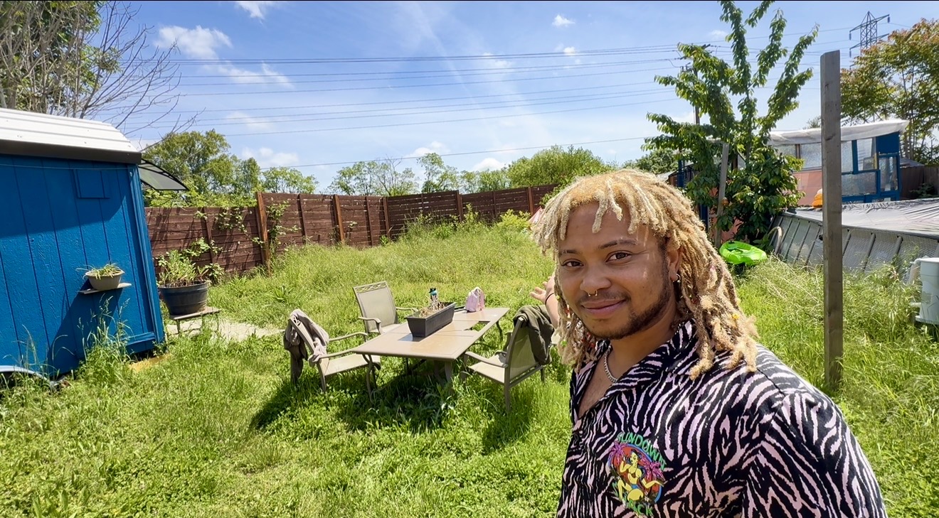 Young person with blonde dreadlocks smiling outdoors in a backyard with a table and chairs, blue shed, and trees.