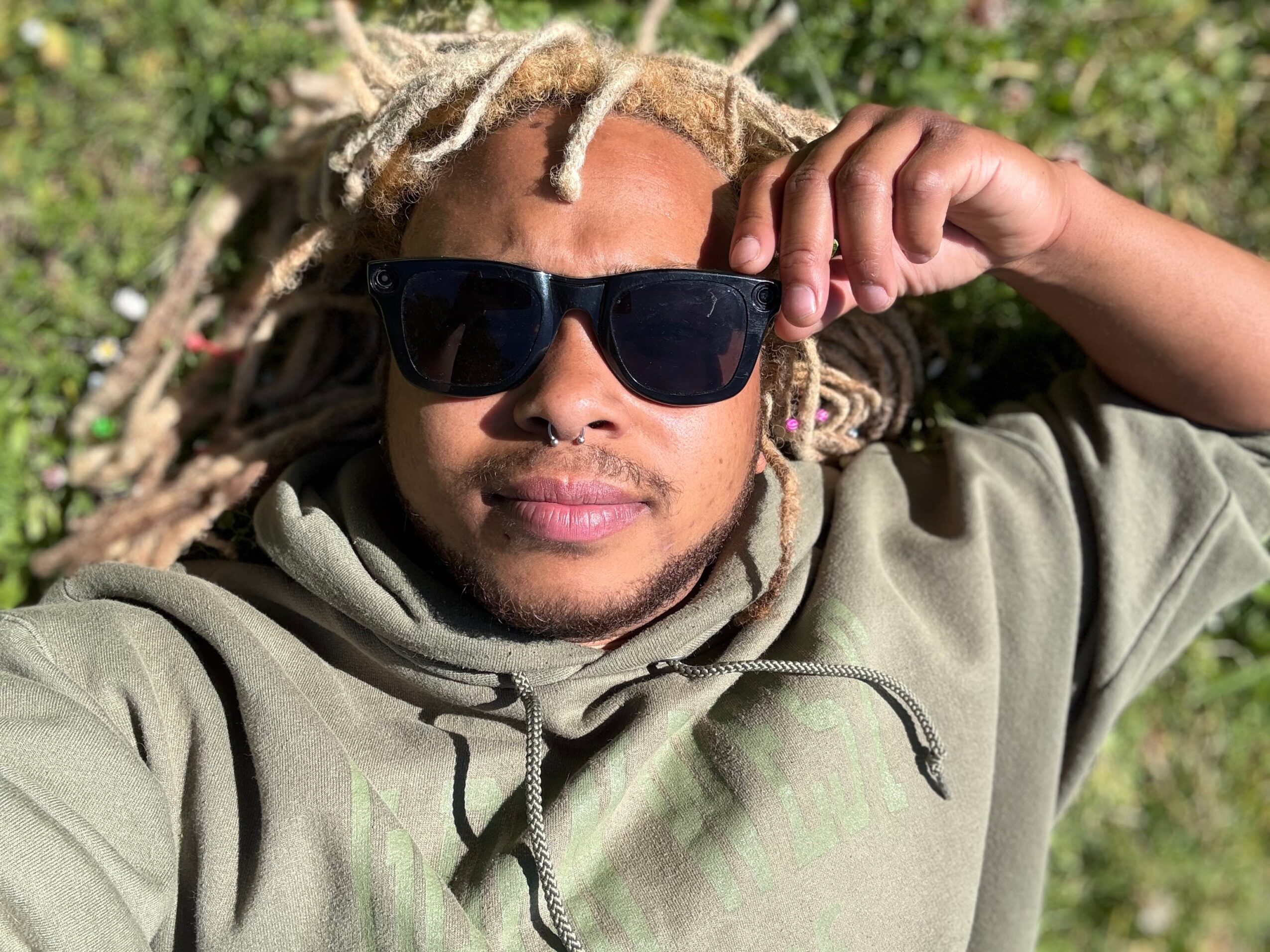 Person with dreadlocks wearing sunglasses and a hoodie, lying outdoors with greenery in the background.
