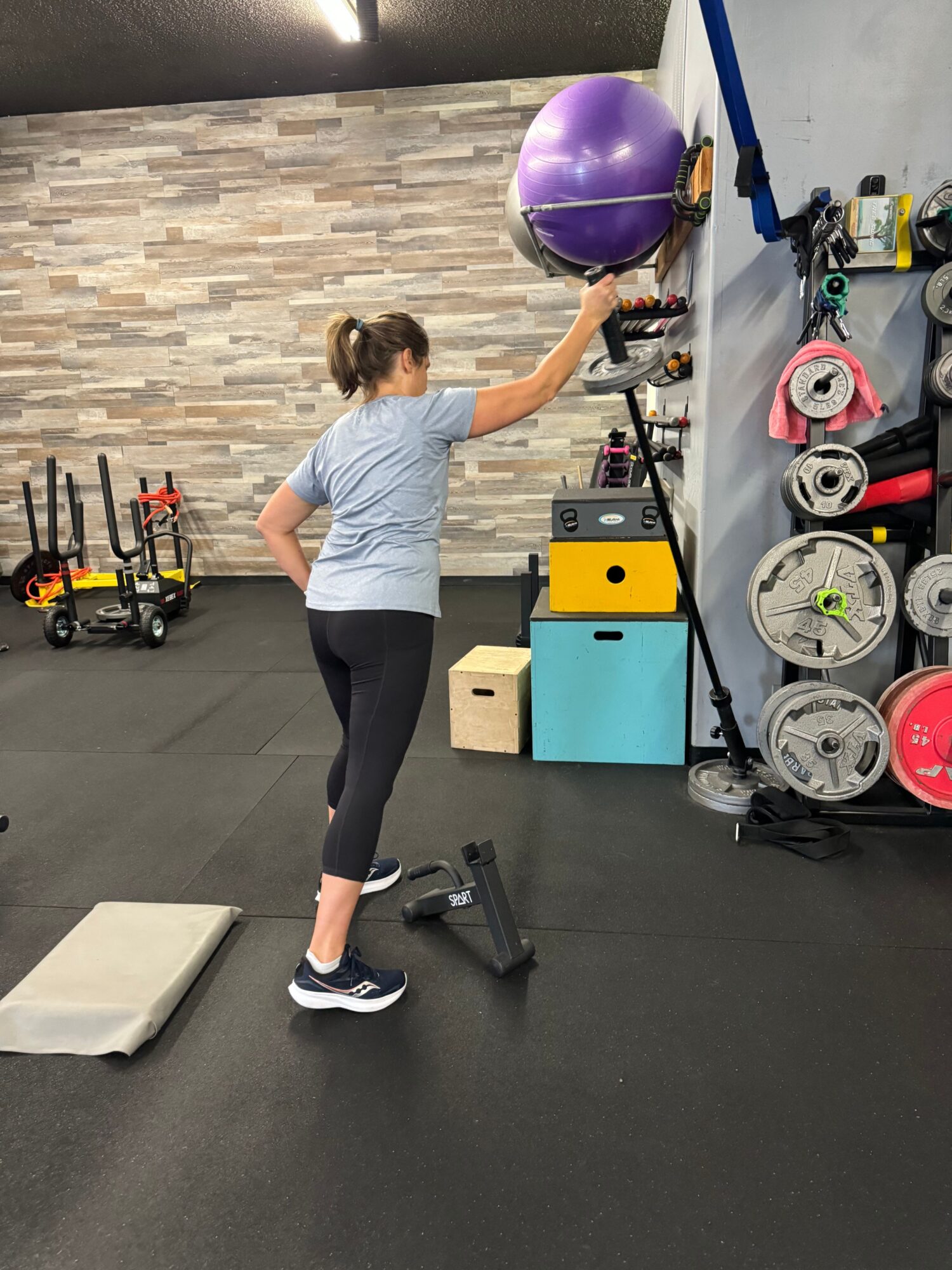 Woman in gym lifting a kettlebell with one arm, gym equipment and weights in background.
