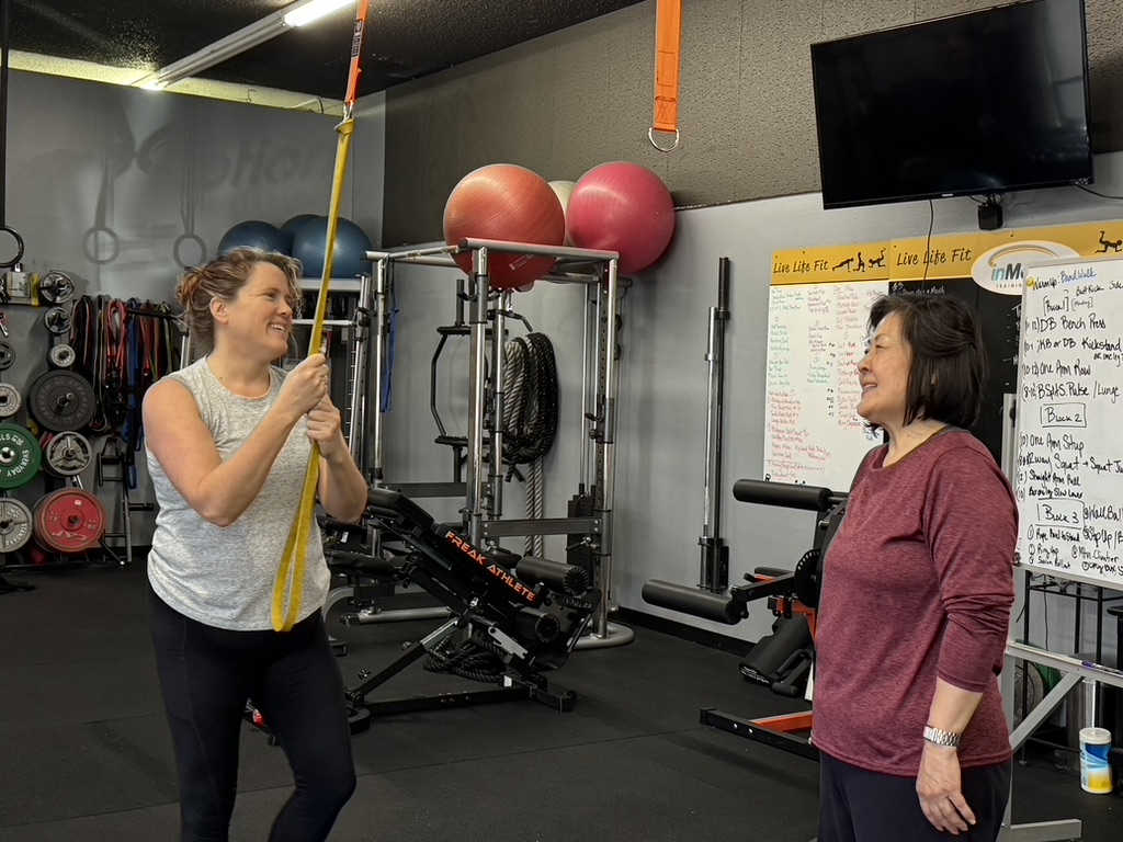 Two women in a gym, one holding a yellow resistance band, smiling and talking, with exercise equipment and a whiteboard nearby.