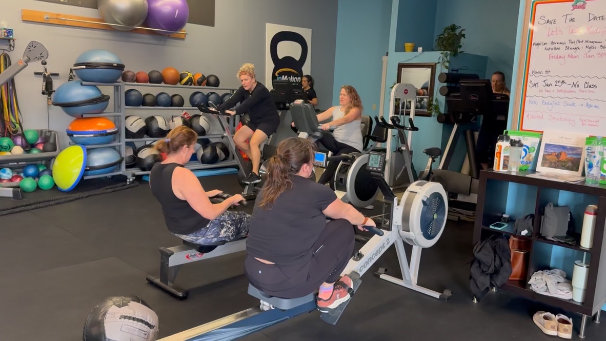 People exercising with rowing machines and weights in a gym, shelves with fitness equipment in the background.