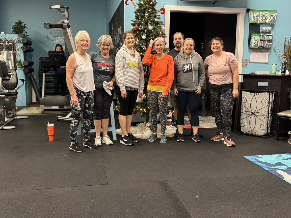 Group of eight people standing in a gym with a Christmas tree, smiling, wearing casual workout clothes.