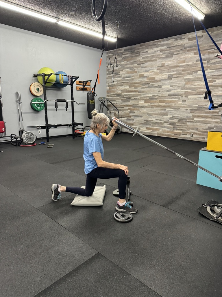 Woman kneeling on a mat in a gym, holding a resistance band, with gym equipment and a brick wall in the background.