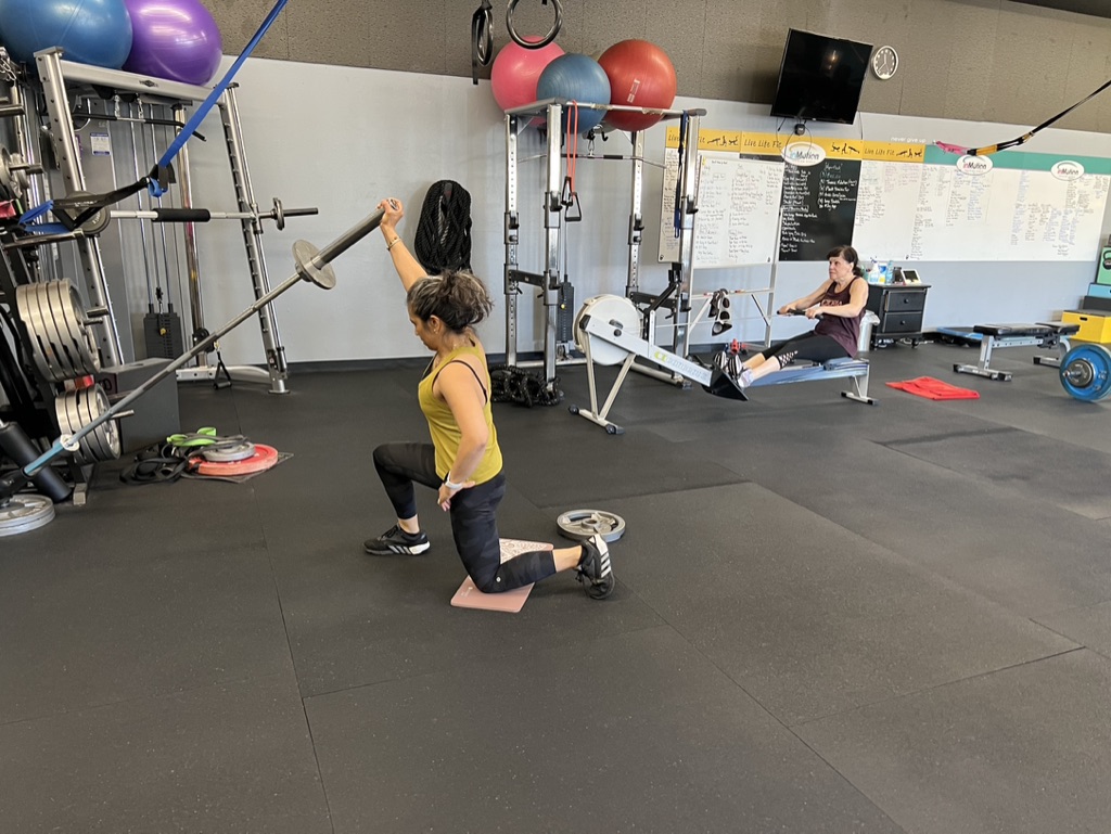 A woman kneels on a mat in a gym, holding a barbell overhead, with a woman sitting on a rowing machine in the background.