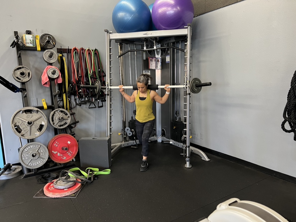 Person in yellow shirt lifting a barbell in a gym with exercise equipment and large exercise balls behind.