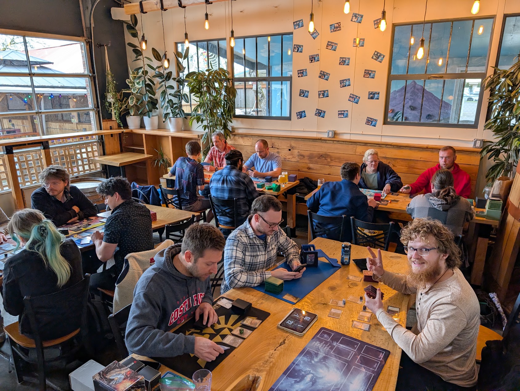 People playing board games at tables in a well-lit cafe with large windows and plants.