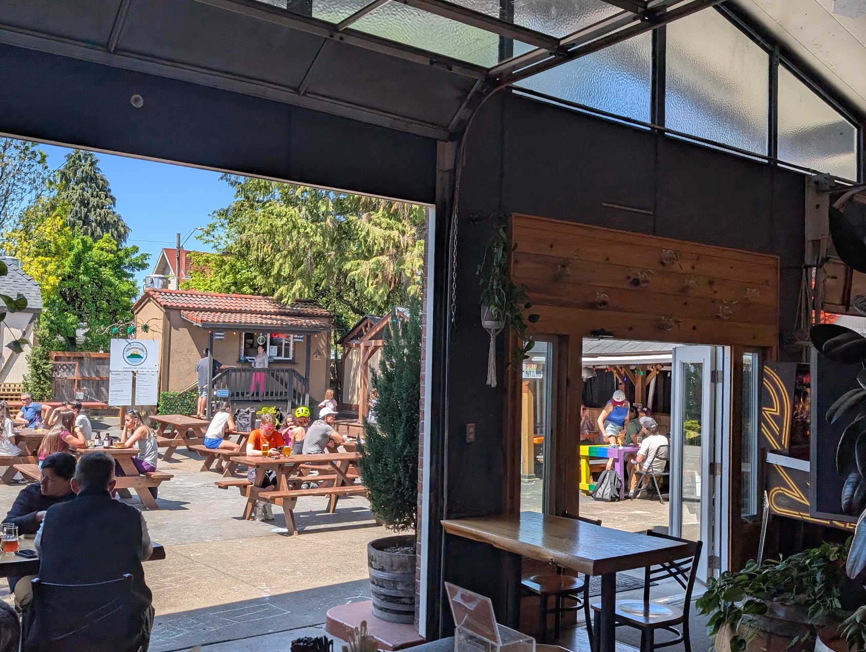 Indoor view looking out to outdoor seating area with people, trees, and buildings, sunlight, and clear sky.
