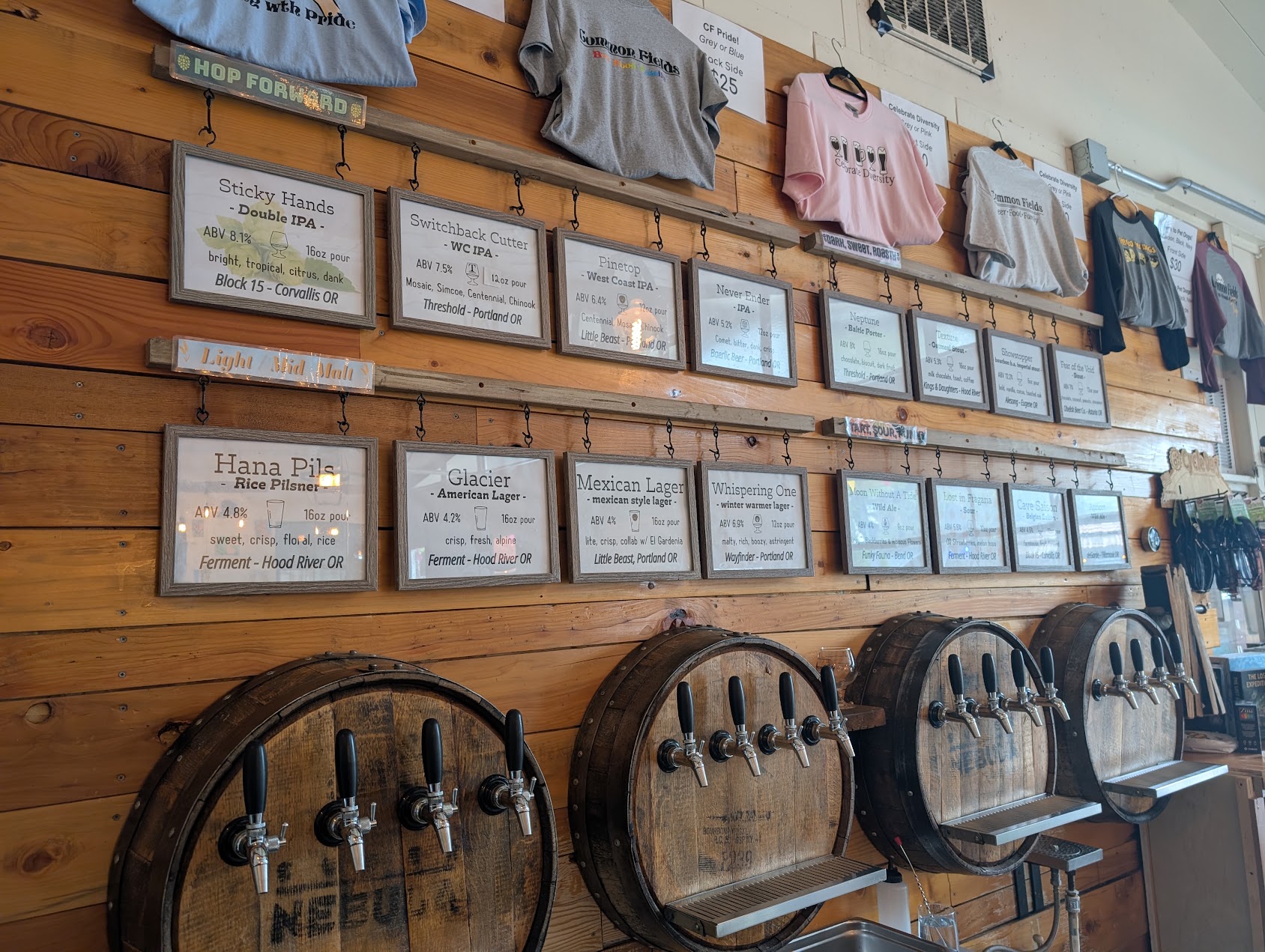 Wooden wall with framed menus, T-shirts hanging above, and beer taps with barrels below.