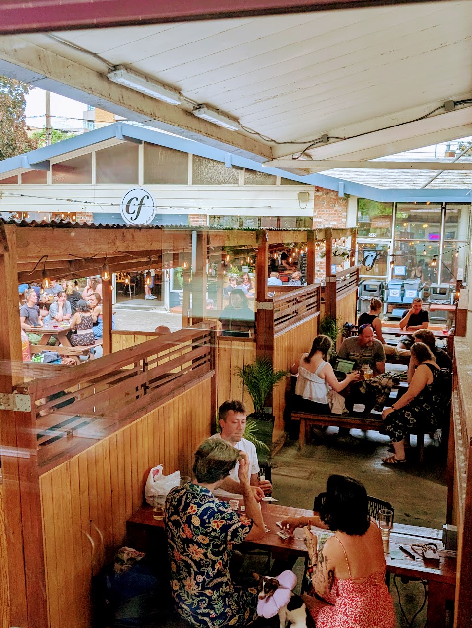 People dining at tables inside a restaurant with large windows and outdoor seating visible through glass walls.