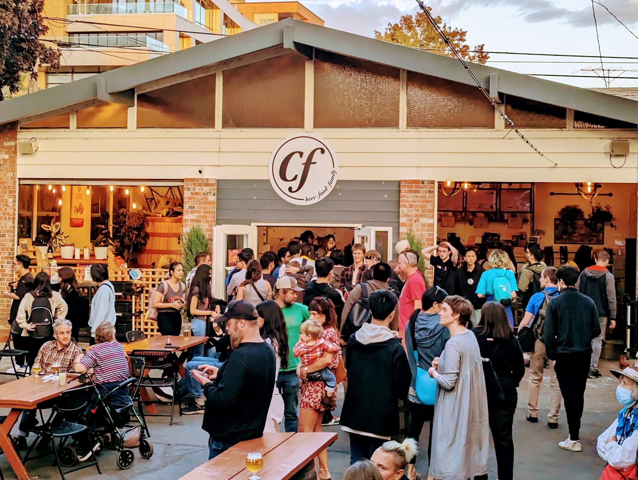 Crowd of people gathered outside a cafe with a circular sign displaying 'cf' logo, some sitting and others standing.