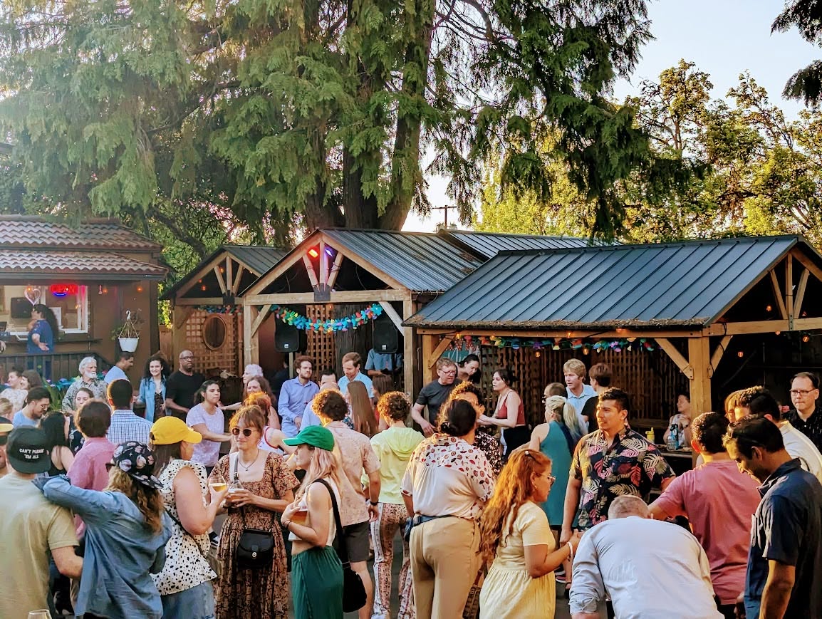 Crowd gathered outdoors near wooden structures decorated with string lights and bunting, with trees and a building in the background.