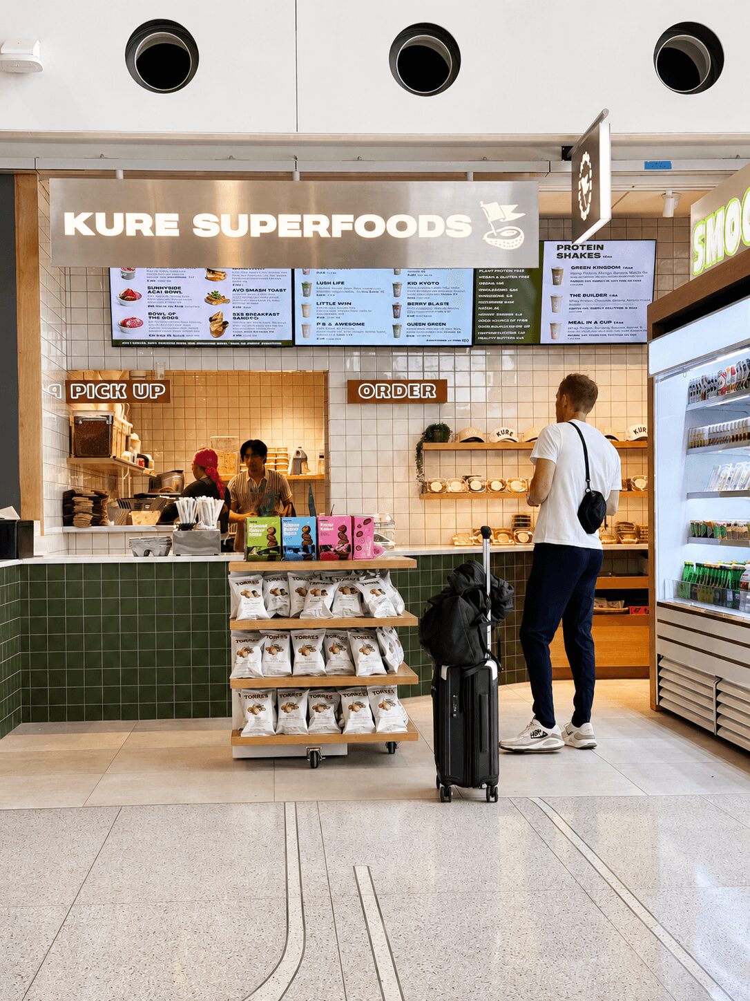 Customer at a food counter with menu boards, shelves with products, and a refrigerated display on the right.