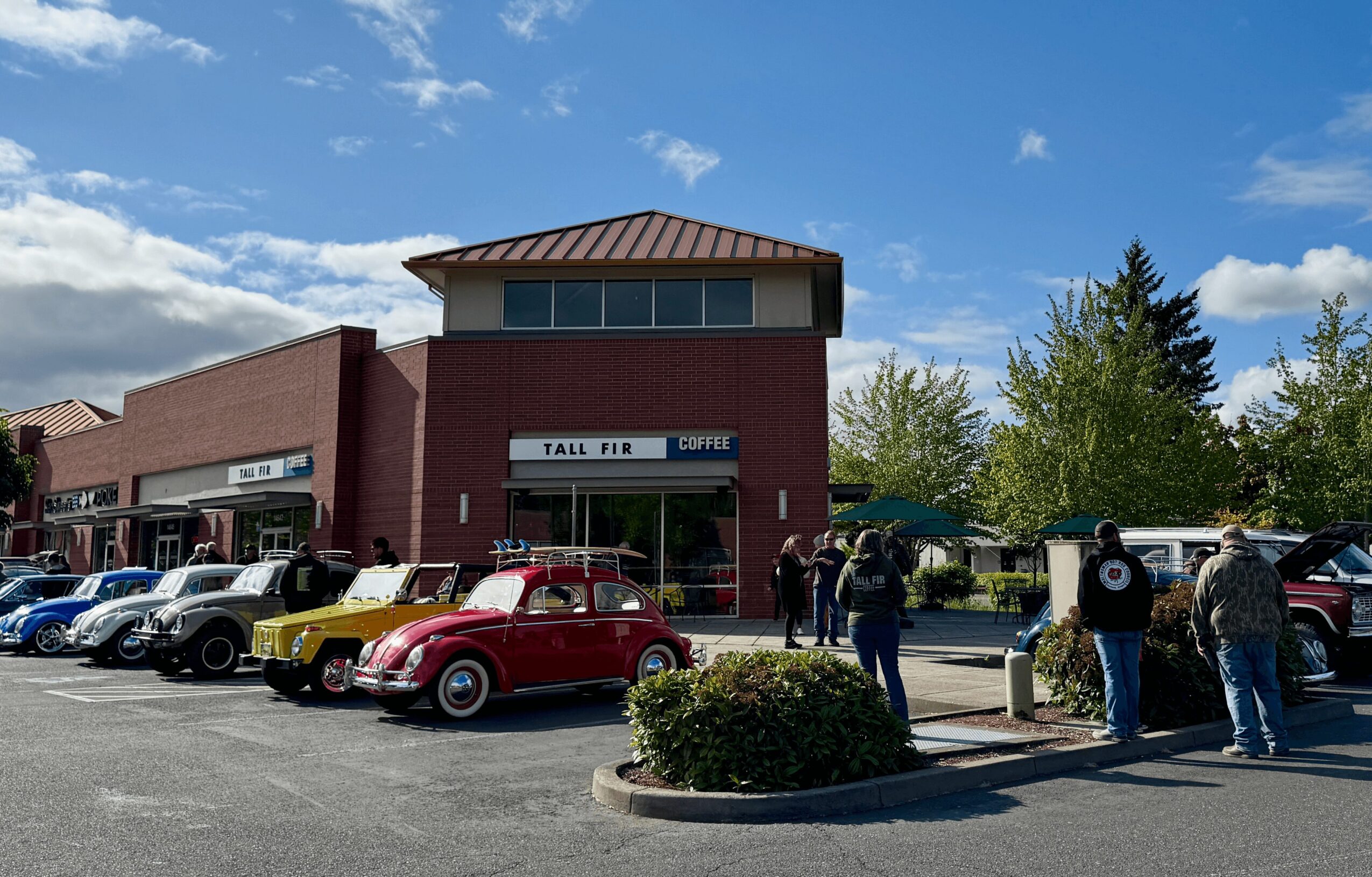 People gather outside a brick building with a parking lot filled with vintage cars, under a blue sky with clouds.