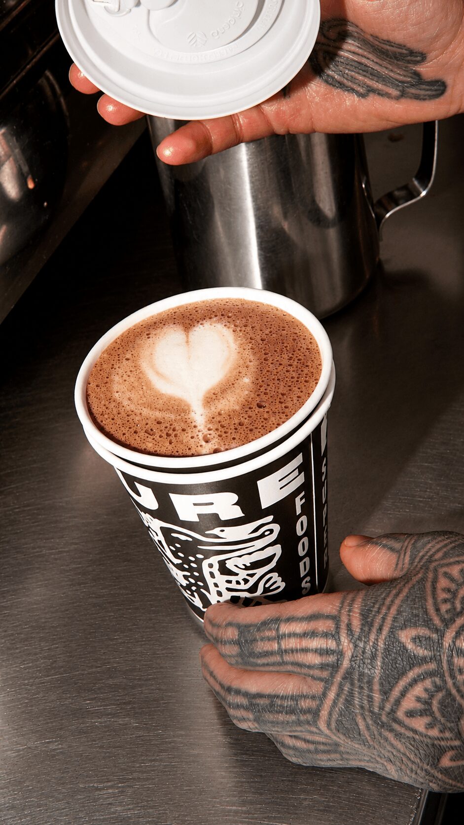 Person with tattoos pouring steamed milk into coffee with heart-shaped latte art, holding a cup with a black and white design