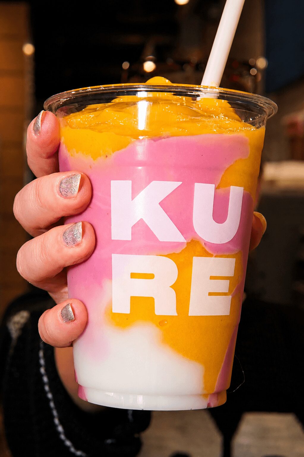 Hand holding a colorful drink with pink, yellow, and white layers in a clear cup with white text, straw inside.