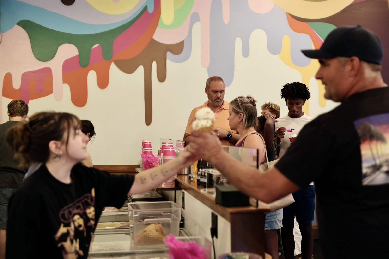 Girl and man exchanging ice cream cones in a colorful ice cream shop with other customers in the background.