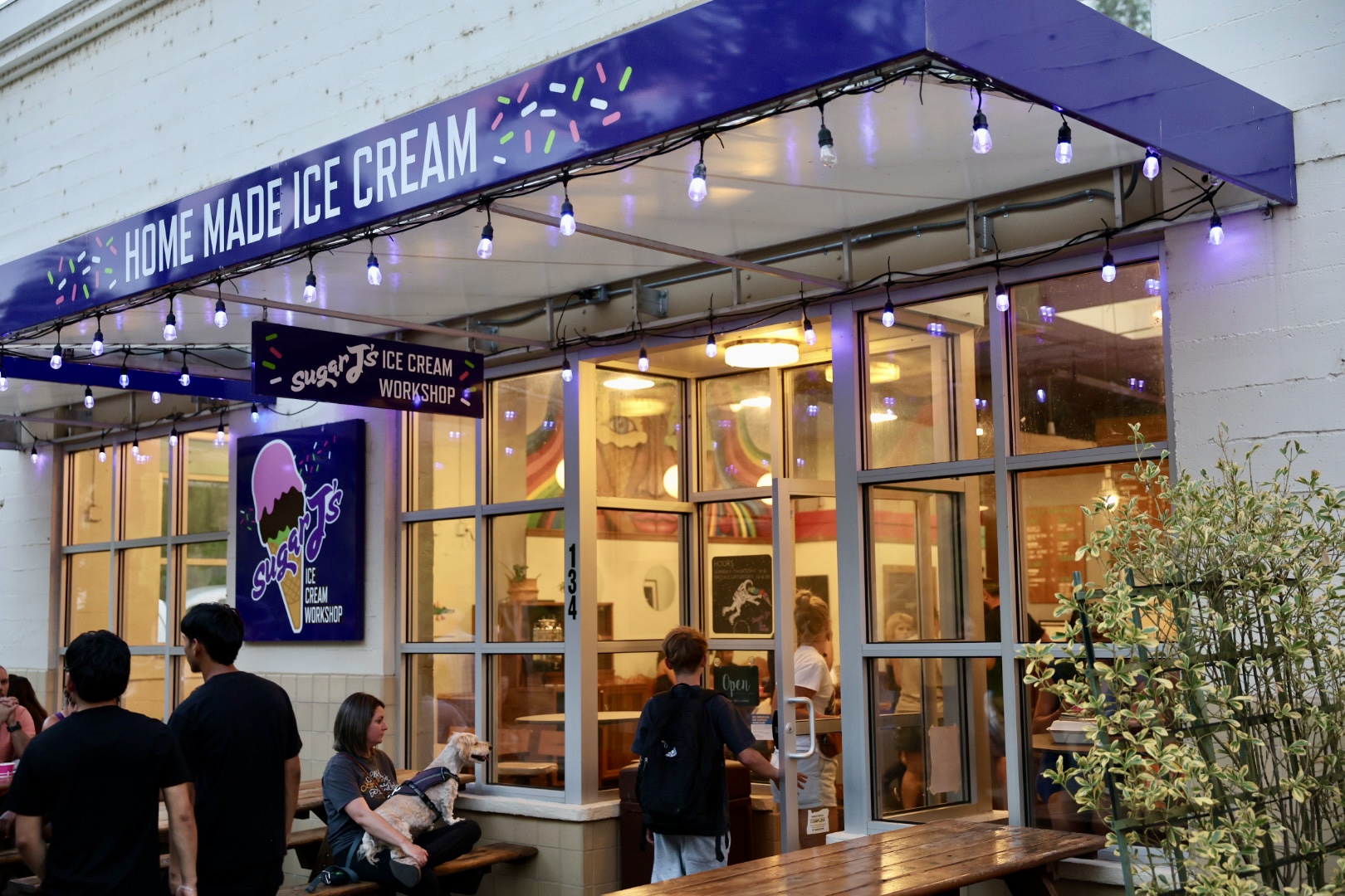 People outside a cream shop with a blue awning and string lights, large glass windows showing the interior, and a sign reading 'Home Made Ice Cream'.