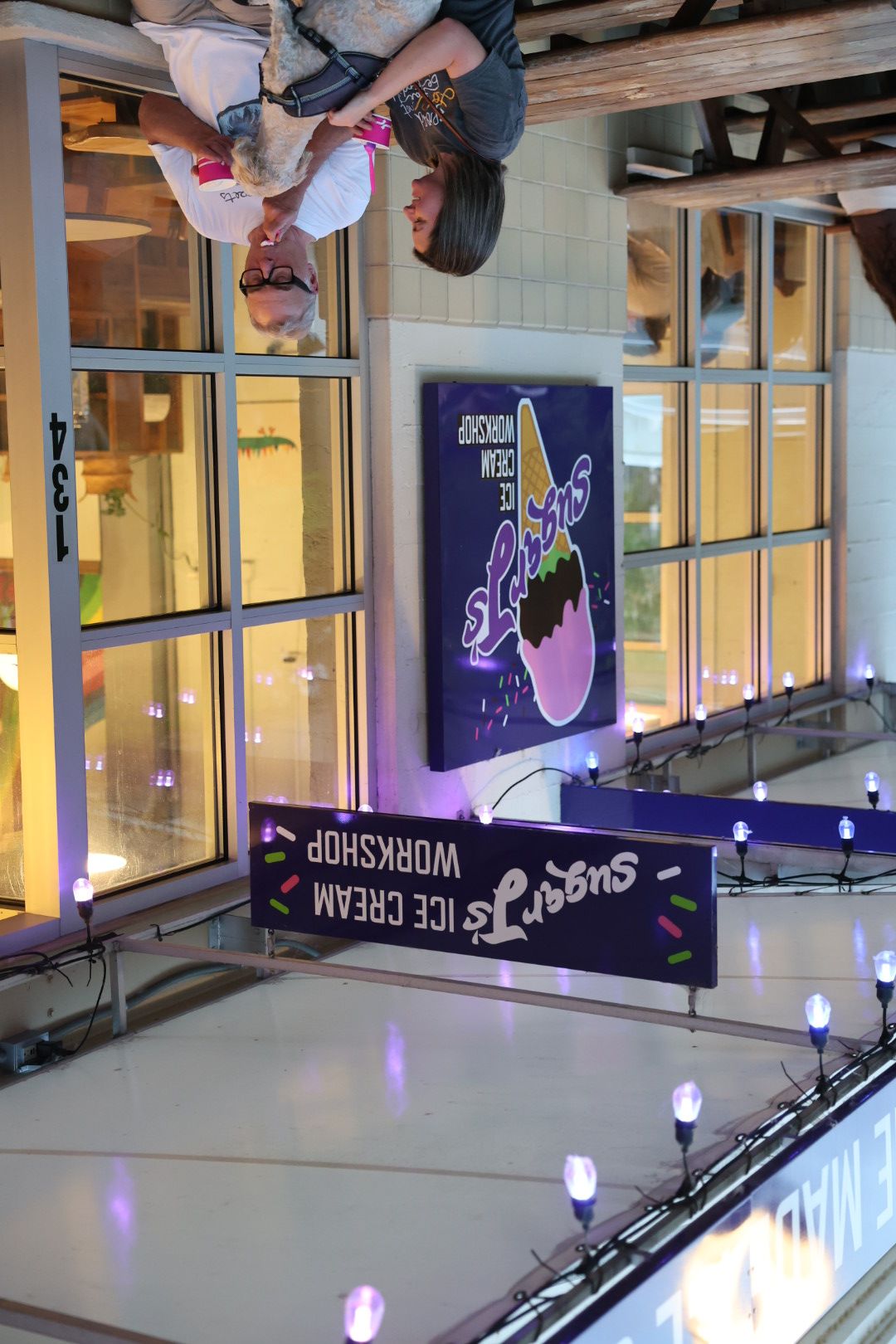 People sitting outside a building with glass windows and a sign for ice cream and workshop, with purple lights overhead.