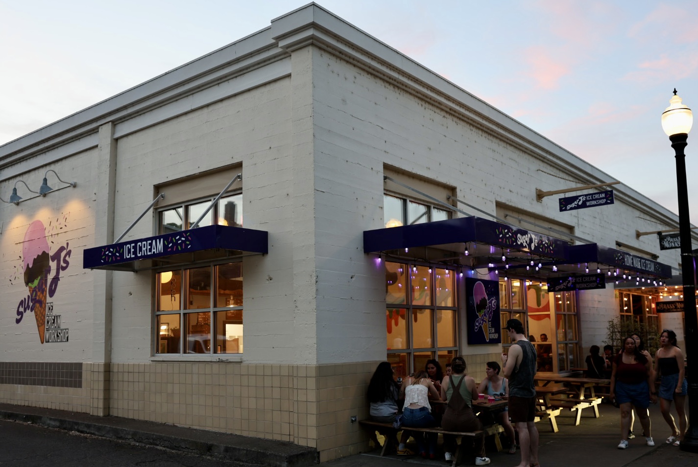 Corner of an ice cream shop with outdoor seating, people sitting and standing outside, and a streetlamp nearby.