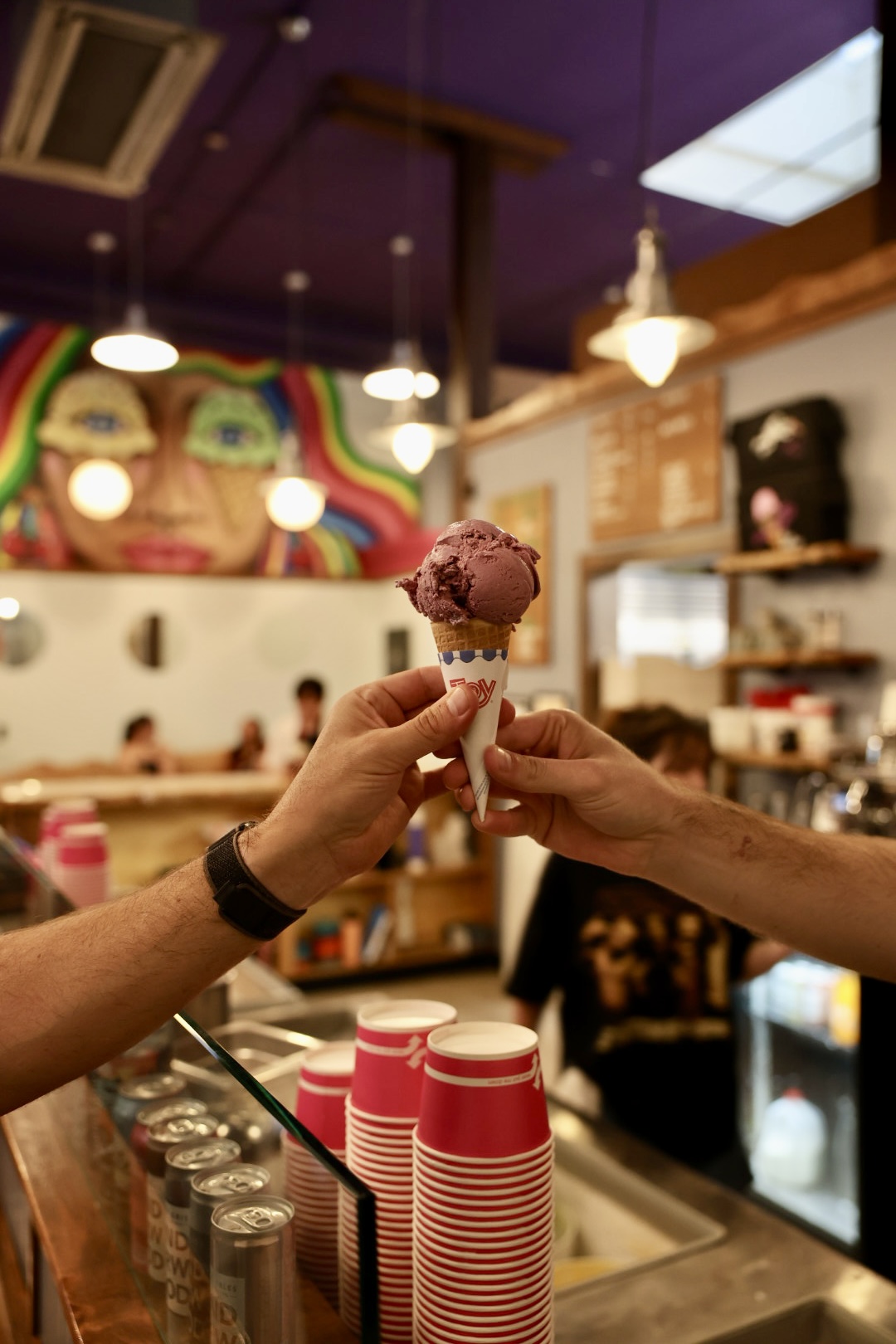 Person hands over a scoop of purple ice cream in a cone to another person inside an ice cream shop, colorful mural in background.