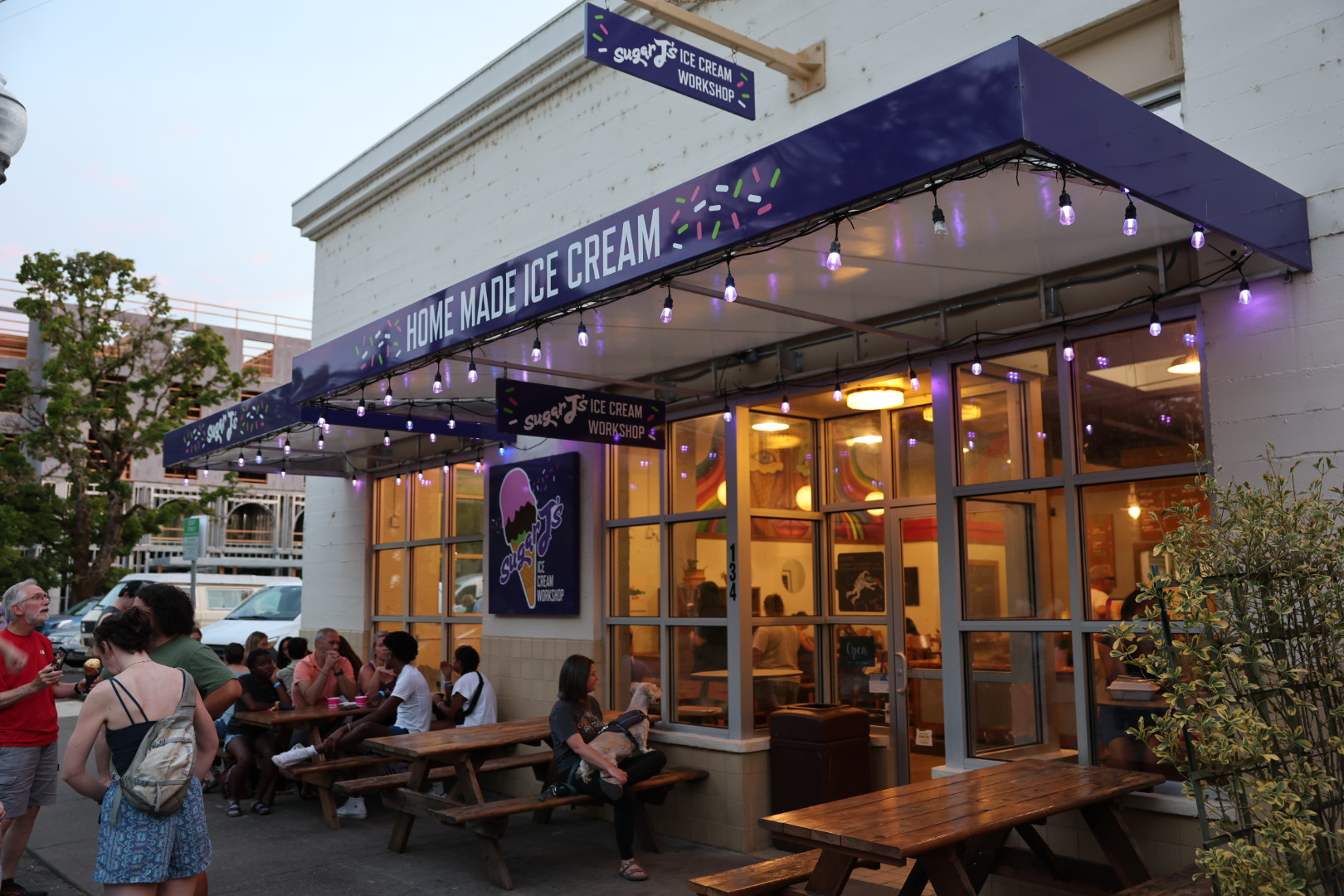 People sitting outside an ice cream shop under a blue awning with string lights, in a lively urban setting.
