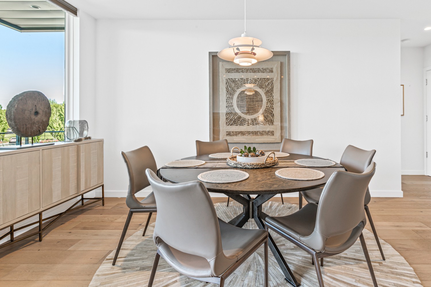 Round dining table with six chairs in a bright room, window with view outside, artwork on wall, wooden floor.