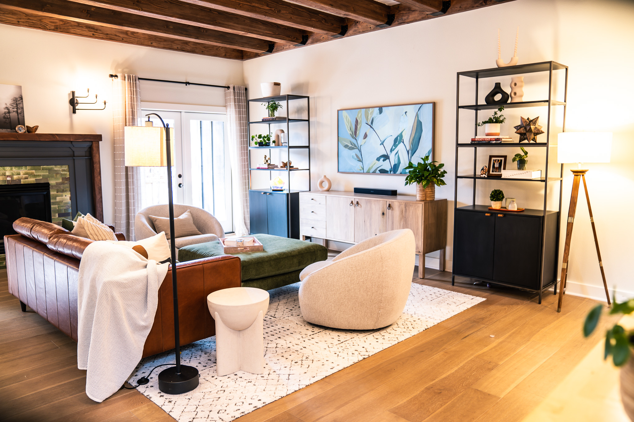 Living room with wooden ceiling beams, sofa, armchair, TV, and shelves, decorated with plants and lamps, hardwood floor, and a rug.